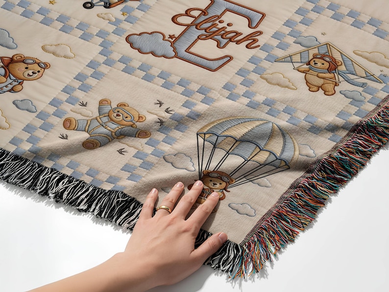 May include: A woven throw blanket featuring a beige background with a blue and white checkered pattern. The design includes illustrations of teddy bears wearing aviator goggles, flying with parachutes, kites, and planes, and the name Elijah.