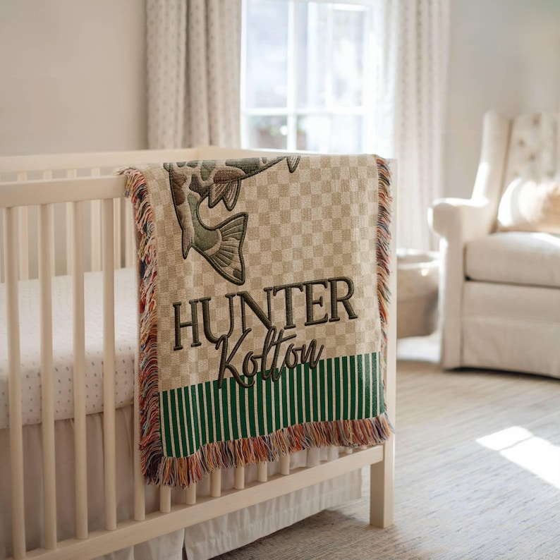 May include: A beige and green baby blanket with a fish design and the name "Hunter Kolton" embroidered in dark green. The blanket has a checkered pattern, green stripes, and a fringed edge. It is draped over a white crib.