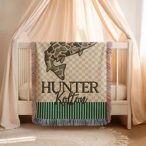 May include: A beige and green baby blanket with a fish design and the name "Hunter Kolton" embroidered on it. The blanket has a checkered pattern and green and white stripes at the bottom, with fringed edges. The blanket is draped over a white crib.