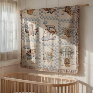 May include: A woven throw blanket featuring a grid pattern with illustrations of teddy bears flying in airplanes, hang gliders, and parachutes. The center panel displays a large letter E with the name Elijah written in cursive script.