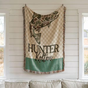 May include: A beige and green woven throw blanket with a large fish design. The fish has a camouflage pattern. The blanket has the name "Hunter Kolton" embroidered on it. The bottom has green and white stripes.