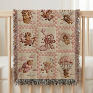 May include: A woven throw blanket featuring a grid pattern with light pink and beige tones. The design displays teddy bears in various aviation scenes, including hot air balloons, aeroplanes, and parachutes. A large letter R with the name Rhea is centred.