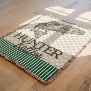 May include: A woven throw blanket with a fish design and the text "HUNTER Kolton". The blanket features a checkered pattern, a green striped border, and a fringed edge. The fish is in shades of green and brown.