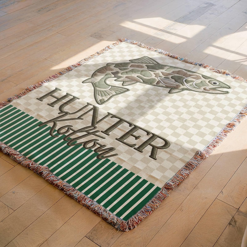 May include: A woven throw blanket with a fish design and the text "HUNTER Kolton". The blanket features a checkered pattern, a green striped border, and a fringed edge. The fish is in shades of green and brown.