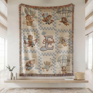 May include: A woven throw blanket featuring a grid of light blue and cream squares. The design displays teddy bears in aviation gear, including hot air balloons, airplanes, parachutes, and jetpacks. The center features the name Elijah in script.