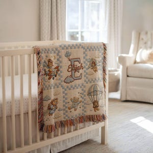 May include: A woven throw blanket featuring a grid pattern with illustrations of teddy bears in aviation gear, including airplanes, parachutes, and jetpacks. The center displays a large letter E with the name Elijah embroidered in brown thread.
