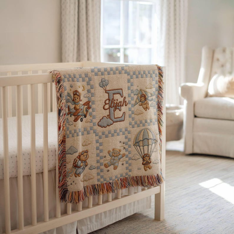May include: A woven throw blanket featuring a grid pattern with illustrations of teddy bears in aviation gear, including airplanes, parachutes, and jetpacks. The center displays a large letter E with the name Elijah embroidered in brown thread.