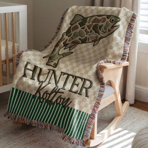 May include: A beige and green checkered throw blanket with a large embroidered fish design. The blanket has the name "Hunter Kolton" embroidered in dark green. The bottom edge features green and white stripes and fringe.