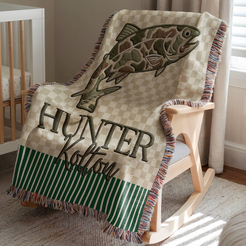 May include: A beige and green checkered throw blanket with a large embroidered fish design. The blanket has the name "Hunter Kolton" embroidered in dark green. The bottom edge features green and white stripes and fringe.