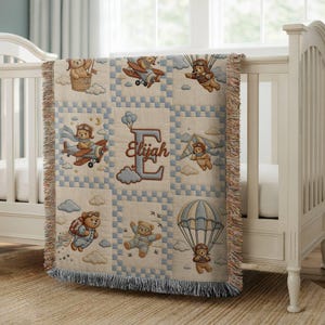 May include: A woven throw blanket featuring a grid pattern with illustrations of teddy bears in various flying modes, including a hot air balloon, airplane, hang glider, jetpack, and parachute, centered with a large letter E and the name Elijah.
