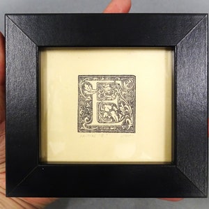 May include: A black framed print of a letter E with ornate floral designs. The print is on a cream background and has the text "Tanner '18" in the bottom left corner.