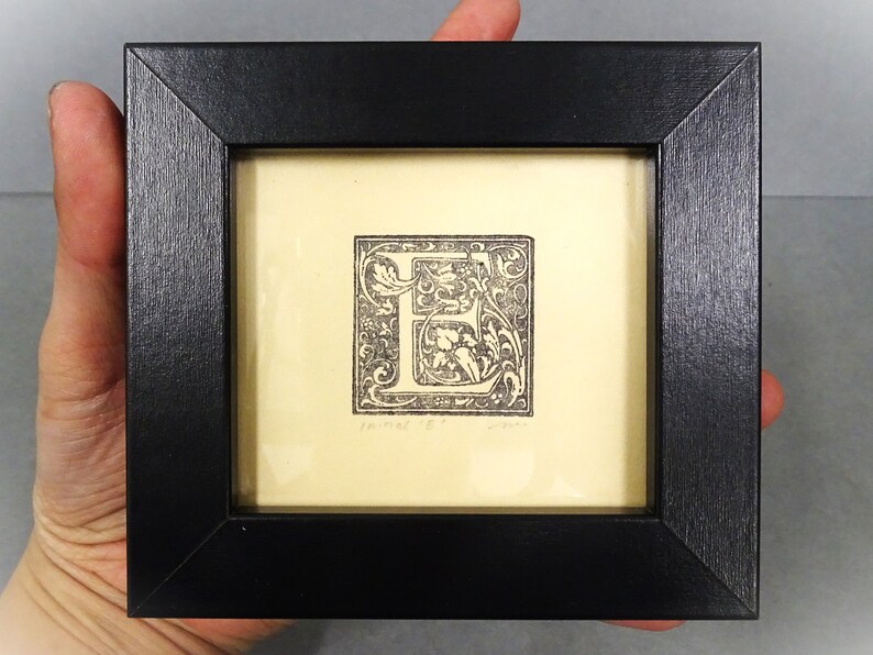 May include: A black framed print of a letter E with ornate floral designs. The print is on a cream background and has the text "Tanner '18" in the bottom left corner.