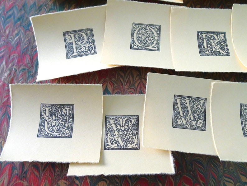 May include: A set of nine cream-colored cards with black ink, each featuring a different letter of the alphabet in a decorative, ornate font.