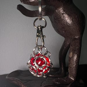 May include: A silver keychain with a red glass ball encased in a metal cage. The keychain is hanging from a frog-shaped metal stand.
