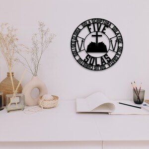 Puede incluir: Arte de pared de metal negro con forma de c&iacute;rculo con las palabras "Five Solas" y "Sola Scriptura", "Sola Fide", "Sola Gratia", "Solus Christus" y "Sola Gloria" alrededor del exterior. El centro del c&iacute;rculo presenta una cruz sobre un libro abierto con una monta&ntilde;a detr&aacute;s.
