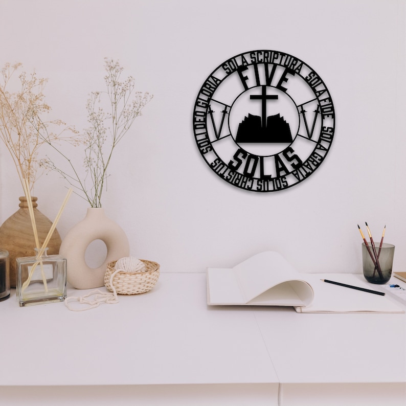 Puede incluir: Arte de pared de metal negro con forma de c&iacute;rculo con las palabras "Five Solas" y "Sola Scriptura", "Sola Fide", "Sola Gratia", "Solus Christus" y "Sola Gloria" alrededor del exterior. El centro del c&iacute;rculo presenta una cruz sobre un libro abierto con una monta&ntilde;a detr&aacute;s.