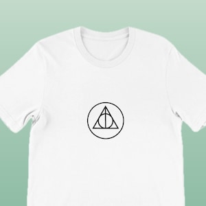 Può includere: T-shirt bianco con grafica nera di un triangolo, una linea verticale e un cerchio. La maglietta ha girocollo e maniche corte. Il disegno è centrato sul petto.
