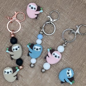May include: Five keychains featuring a sloth design. Each keychain has a different colored sloth and beads. The keychains are made of metal and plastic.