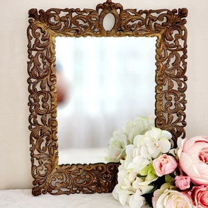 May include: A gold-toned ornate mirror with a decorative, intricate frame. The frame is made of metal and has a floral design. The mirror is reflecting a bouquet of white and pink flowers.