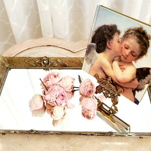 May include: A gold-toned metal tray with a mirrored surface and ornate floral details. The tray features a decorative stand holding a framed print of two cherubs kissing. A bouquet of pink peonies is placed on the tray.