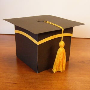 May include: A black graduation cap box with a yellow tassel. The box is decorated with a yellow band.