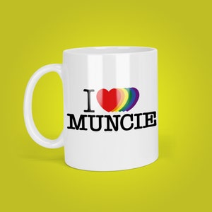 May include: White ceramic coffee mug with a black graphic that says "I love Muncie" with a rainbow heart.