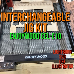 May include: A black and gray laser cutter with a piece of wood on the cutting bed. The wood has a design cut into it that says "Interchangeable Jig Kit" and "Enjoywood Cel-E10". The laser cutter has a red laser head and a black and gray control panel. The text "The Clack Shack" is visible on the side of the laser cutter.