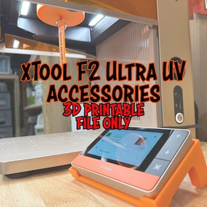 Puede incluir: Un panel de control XTool naranja y plateado con pantalla táctil, sobre un soporte naranja. La imagen muestra una impresora 3D con el texto "XTool F2 Ultra UV Accessories 3D Printable File Only."