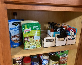 May include: A kitchen cabinet shelf with various food items and a wooden organizer. The organizer has three compartments and is labeled "Clip-all". The food items include pasta, trail mix, and hot sauce.
