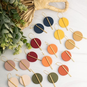 May include: A collection of circular earrings in various colors, including navy, yellow, red, orange, tan, pink, and olive green. Each earring features a textured round disc attached to a thin gold-tone metal hoop and a vertical gold-tone metal bar.