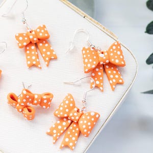 May include: Orange bow earrings with white polka dots. The earrings are made of a smooth material and have a silver hook. The bows are a bright orange color and are arranged on a white surface.