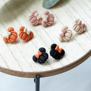 May include: A set of six pairs of polymer clay earrings. The earrings are shaped like pumpkins and Mickey Mouse heads. The pumpkins are orange and the Mickey Mouse heads are black with orange bows. The earrings are displayed on a wooden stand.