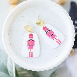 Puede incluir: Pendientes colgantes con un diseño de cascanueces rosa sobre fondo blanco. Los pendientes tienen tachuelas doradas y un acento ovalado dorado. Los pendientes se exhiben en un plato blanco moteado.