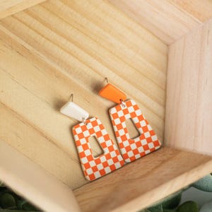 K&ouml;nnte beinhalten: Ein Paar orange-wei&szlig; karierte Ohrringe mit rechteckiger Form und einem Ausschnitt in der Mitte. Die Ohrringe haben ein wei&szlig;es Oberteil und ein orangefarbenes Oberteil. Die Ohrringe werden in einer Holzkiste pr&auml;sentiert.