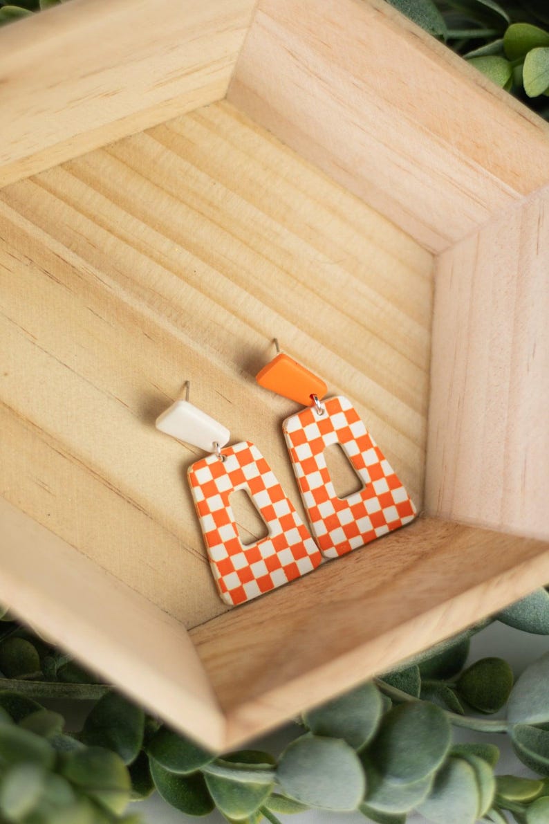 K&ouml;nnte beinhalten: Ein Paar orange-wei&szlig; karierte Ohrringe mit rechteckiger Form und einem Ausschnitt in der Mitte. Die Ohrringe haben ein wei&szlig;es Oberteil und ein orangefarbenes Oberteil. Die Ohrringe werden in einer Holzkiste pr&auml;sentiert.