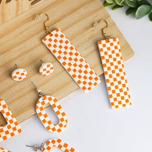 K&ouml;nnte beinhalten: Eine Sammlung orange-wei&szlig; karierter Ohrringe in verschiedenen Formen, darunter lange Rechtecke, Ovale und kleine Ohrstecker. Die Ohrringe werden auf einer Holzoberfl&auml;che mit wei&szlig;em Hintergrund pr&auml;sentiert und zeigen ein modernes Schmuckdesign.