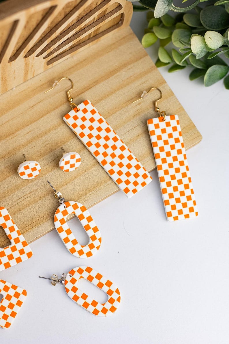 K&ouml;nnte beinhalten: Eine Sammlung orange-wei&szlig; karierter Ohrringe in verschiedenen Formen, darunter lange Rechtecke, Ovale und kleine Ohrstecker. Die Ohrringe werden auf einer Holzoberfl&auml;che mit wei&szlig;em Hintergrund pr&auml;sentiert und zeigen ein modernes Schmuckdesign.