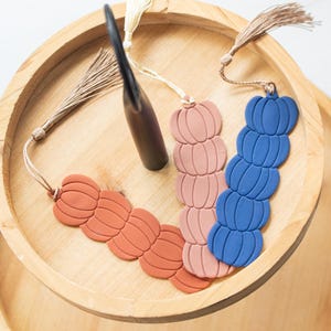 May include: Three fall-themed bookmarks with a pumpkin design. The bookmarks are made of clay and have a tassel. The bookmarks are in shades of orange, pink, and blue.