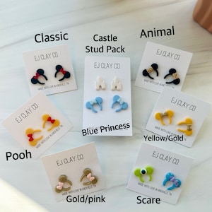 May include: A set of 10 pairs of handmade polymer clay earrings. The earrings are shaped like Mickey Mouse ears and feature various colors and designs, including classic red and black, blue and white, yellow and gold, and gold and pink. The earrings are labeled with the names "Classic", "Castle Stud Pack", "Animal", "Blue Princess", "Yellow/Gold", "Gold/pink", and "Scare".