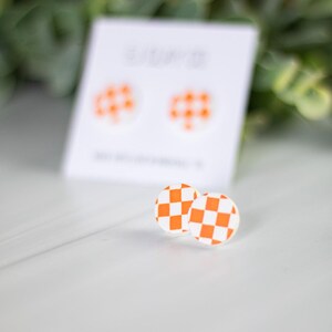 K&ouml;nnte beinhalten: Ein Paar runde Ohrringe mit orange-wei&szlig;em Schachbrettmuster. Die Ohrringe liegen auf einer wei&szlig;en Oberfl&auml;che, mit einem verschwommenen Hintergrund aus Gr&uuml;n. Eine Karte mit passenden Ohrringen befindet sich im Hintergrund.