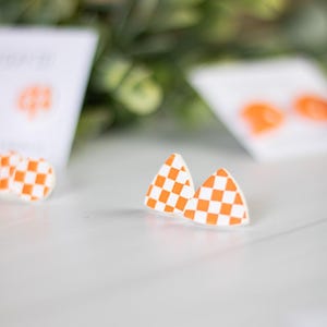 K&ouml;nnte beinhalten: Dreieckige Ohrringe mit wei&szlig;-orangem Schachbrettmuster. Die Ohrringe liegen auf einer wei&szlig;en Oberfl&auml;che, mit einem verschwommenen Hintergrund aus Gr&uuml;n und wei&szlig;en Karten mit orangefarbenen Akzenten. Die Ohrringe haben ein auff&auml;lliges, geometrisches Design.