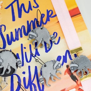 May include: A collection of grey raccoon-shaped accessories, including dangling earrings with black wire hooks, a circular badge reel, and a pink elastic strap with a raccoon charm, all featuring a textured finish and black eye masks.