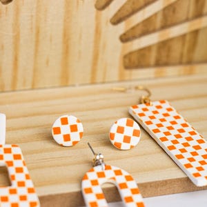 K&ouml;nnte beinhalten: Eine Auswahl an wei&szlig;-orangen karierten Ohrringen in verschiedenen Formen, darunter Stecker, rechteckige H&auml;nger und offene Designs. Die Ohrringe werden auf einer Holzoberfl&auml;che mit einem dekorativen Holz-Hintergrund pr&auml;sentiert.