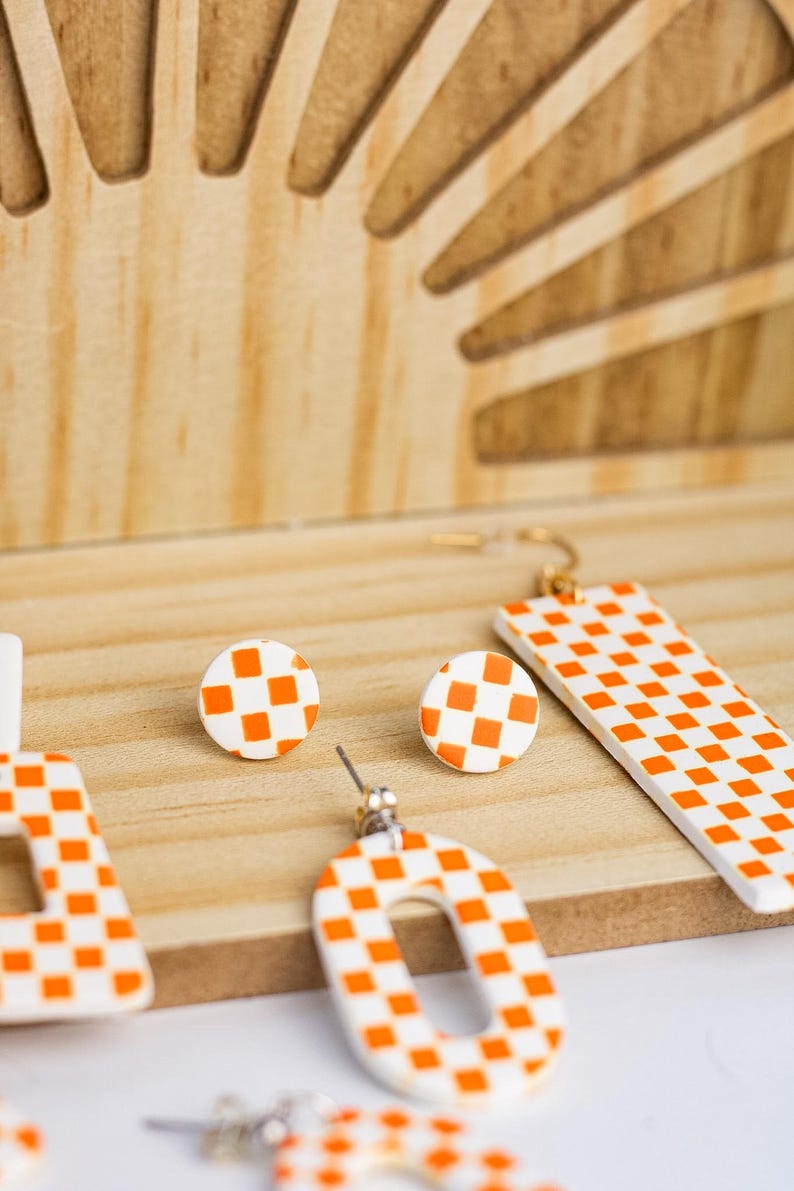 K&ouml;nnte beinhalten: Eine Auswahl an wei&szlig;-orangen karierten Ohrringen in verschiedenen Formen, darunter Stecker, rechteckige H&auml;nger und offene Designs. Die Ohrringe werden auf einer Holzoberfl&auml;che mit einem dekorativen Holz-Hintergrund pr&auml;sentiert.