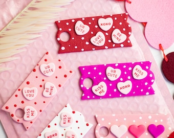 May include: Four pink and red Valentine's Day gift tags with white polka dots and candy heart embellishments. The tags have sayings like "Love You", "Kiss Me", "Be Mine", "Ur Cute", and "XOXO".