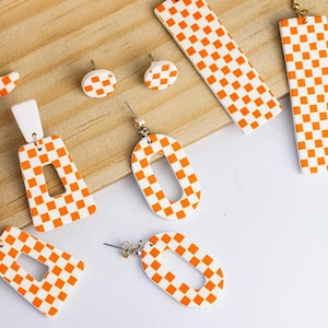 K&ouml;nnte beinhalten: Eine Sammlung handgefertigter Ohrringe in verschiedenen Formen mit einem wei&szlig;-orangen Karomuster. Die Ohrringe umfassen Stecker, rechteckige und ovale Designs, alle mit goldfarbenen Haken. Die Ohrringe werden auf einer Holzoberfl&auml;che pr&auml;sentiert.