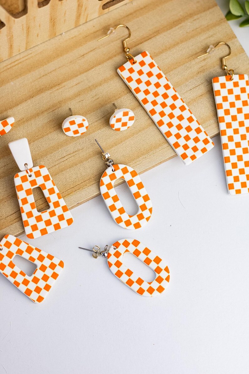 K&ouml;nnte beinhalten: Eine Sammlung handgefertigter Ohrringe in verschiedenen Formen mit einem wei&szlig;-orangen Karomuster. Die Ohrringe umfassen Stecker, rechteckige und ovale Designs, alle mit goldfarbenen Haken. Die Ohrringe werden auf einer Holzoberfl&auml;che pr&auml;sentiert.