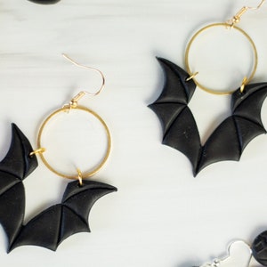 Op de afbeelding: Een paar zwarte vleermuisvormige oorbellen met gouden hoepels. De oorbellen zijn gemaakt van polymer clay en hebben een gedetailleerd ontwerp.