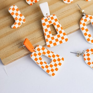 K&ouml;nnte beinhalten: Eine Sammlung handgefertigter Ohrringe und Charms in einem lebendigen orange-wei&szlig;en Schachbrettmuster. Die Ohrringe umfassen rechteckige H&auml;ngeohrringe, ein Paar Creolen und Ohrstecker. Die Charms sind wie Cowboy-Stiefel geformt.