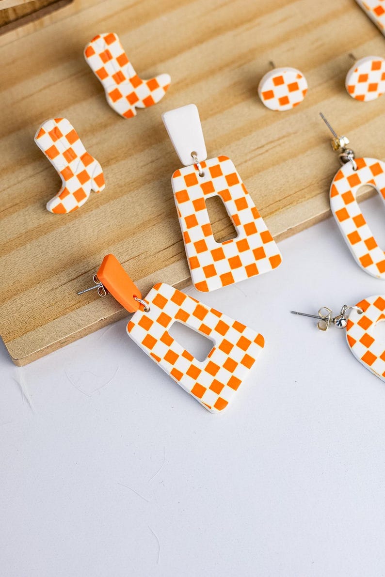 K&ouml;nnte beinhalten: Eine Sammlung handgefertigter Ohrringe und Charms in einem lebendigen orange-wei&szlig;en Schachbrettmuster. Die Ohrringe umfassen rechteckige H&auml;ngeohrringe, ein Paar Creolen und Ohrstecker. Die Charms sind wie Cowboy-Stiefel geformt.